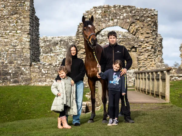 A mum, dad, and their two daughters stand on either side of a large brown horse, smiling for the photo.