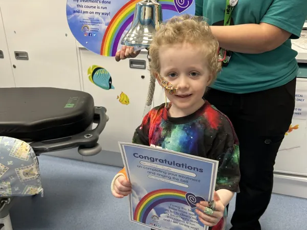 A young boy with short curly blond hair holds a ‘Congratulations’ certificate while standing beside the ‘End of Treatment Bell.’