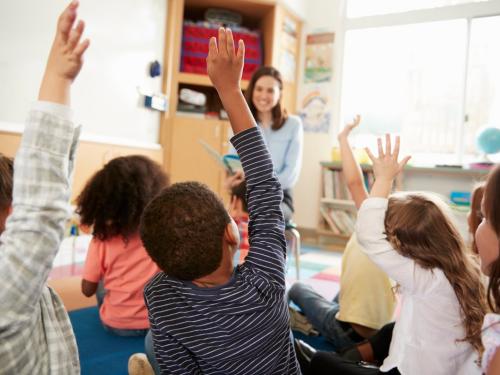 A group of children with their hands in the air, sitting in front of their teacher.