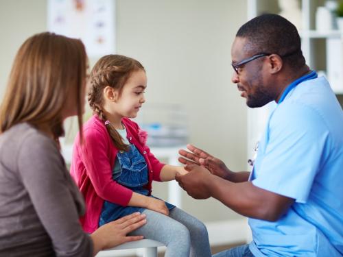 A doctor in blue scrubs talking to a young girl and her mother in a hospital room.