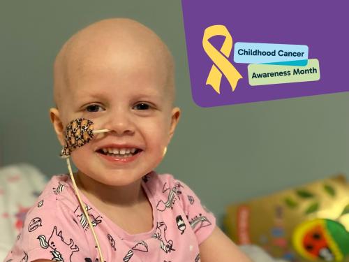 A young girl in pink pyjamas smiles at the camera. She has lost her hair after cancer treatment, and she has a tube in her nose, taped to the side of her face.