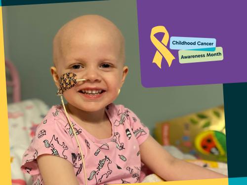A young girl in pink pyjamas smiles at the camera. She has lost her hair after cancer treatment, and she has a tube in her nose, taped to the side of her face.