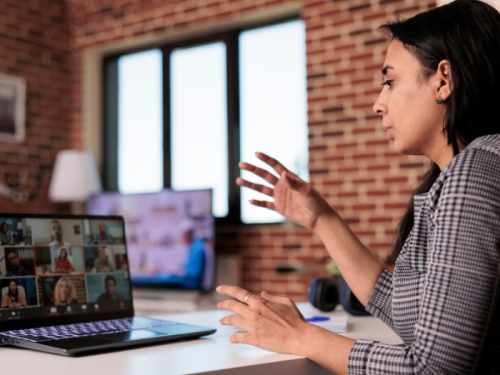 An Indian woman with black hair sitting at a desk with an open laptop on her desk and talking while in an online meeting 