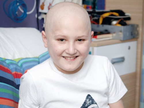 little boy with cancer and bald head sitting on hospital bed reading but looking at camera and smiling