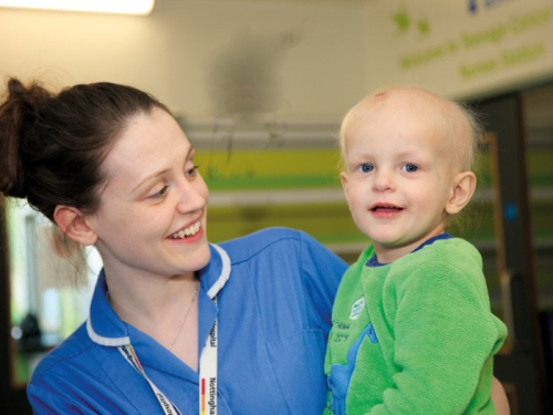 a white nurse in blue uniform in hospital holding a toddler with a green jumper 