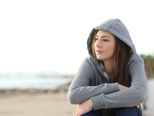 Young adult girl sitting on beach with arms crossed over her knees wearing grey hoodie and looking out contemplatively 