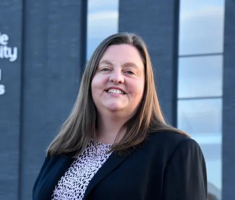 Vikki smiling at the camera and with long straight brown hair and wearing a black jacket.