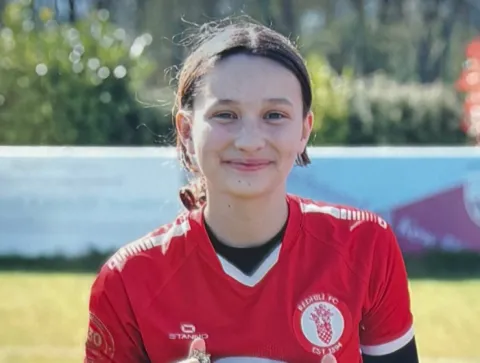 A young girl in her red football kit smiling at the camera in a park