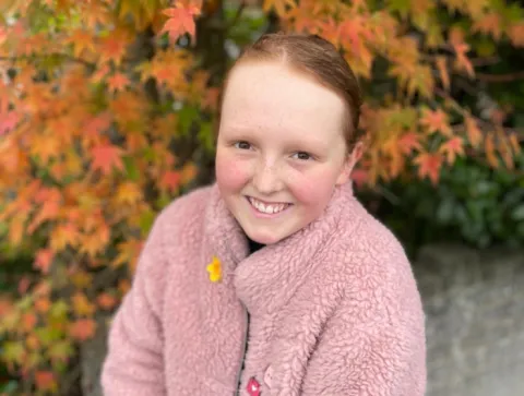 Young girl smiling at camera with pink coat and ginger hair standing in a park with autumn leaves in the bakground