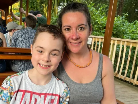 A boy wearing a marvel t shirt and his mum wearing a grey vest and orange necklace sitting in a train