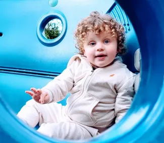 A smiling young boy with curly brown hair is sitting inside a blue climbing frame, wearing a grey tracksuit that matches.