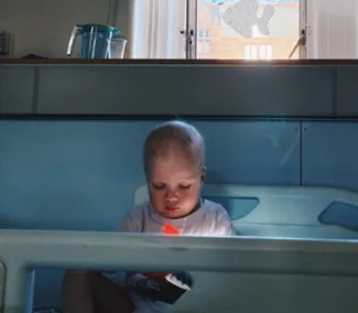 A toddler wearing a light blue t-shirt sits in a hospital bed, with his head down.