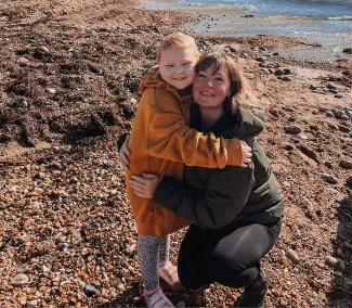 A woman with brown hair, dressed in a dark green coat, is crouched down on the beach, hugging her young daughter. The little girl has short hair and is wearing a bright yellow coat.