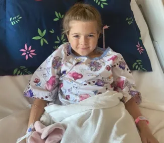 A young girl with brown hair styled in a braid lies in a hospital bed, smiling.