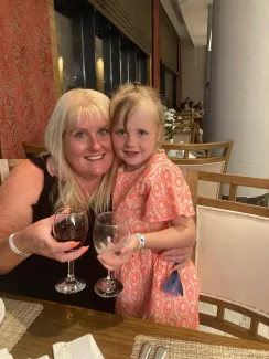 A mother and young daughter sit closely at a dinner table, arms around each other, smiling and holding drinks.