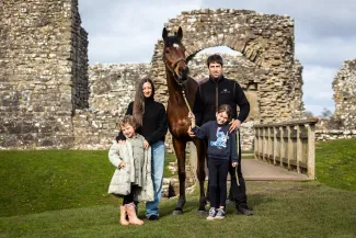 A mum, dad, and their two daughters stand on either side of a large brown horse, smiling for the photo.