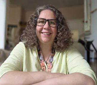 A woman with curly brown hair, glasses, and a yellow top sits with her arms crossed, smiling at the camera.