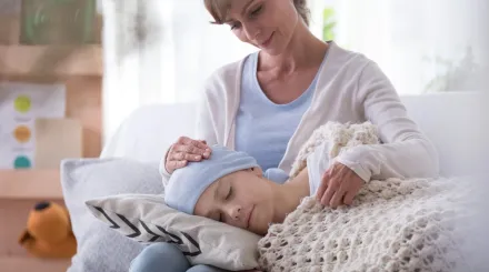 A mother sits on the sofa, with her sick child's head on a cushion in her lap.