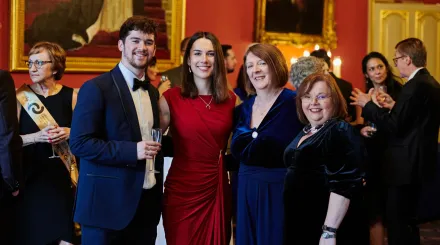 A group of people, one man and three women, dressed in black tie attire in a large event room, smiling at the camera