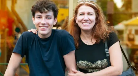 A mother with her arm around her teenage son, both smiling at the camera