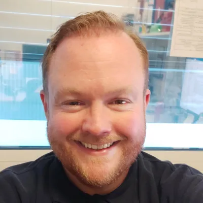 Tom smiling at the camera with short ginger hair and wearing a black t-shirt.
