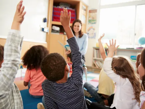 A group of children with their hands in the air, sitting in front of their teacher.