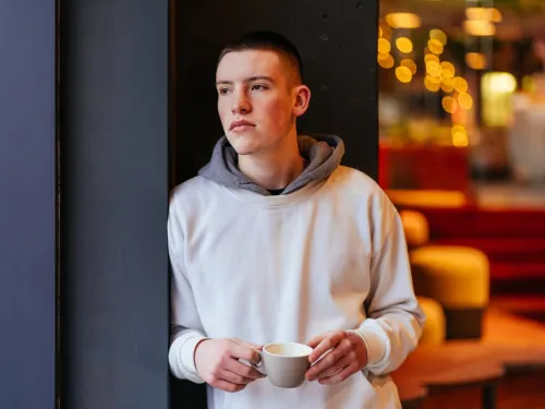 A young man stands in a doorway with a coffee cup, looking thoughtful.
