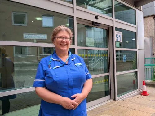 Helen wearing a blue nurses uniform and smiling outside the doors of the hospital.