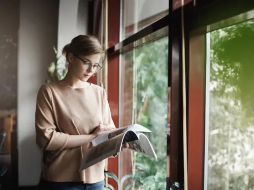 A young woman with glasses, standing near a window, reading a magazine