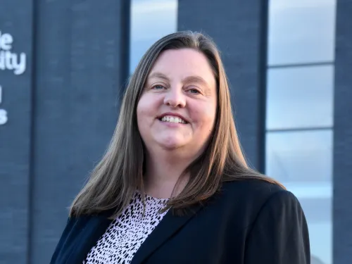Vikki smiling at the camera and with long straight brown hair and wearing a black jacket.