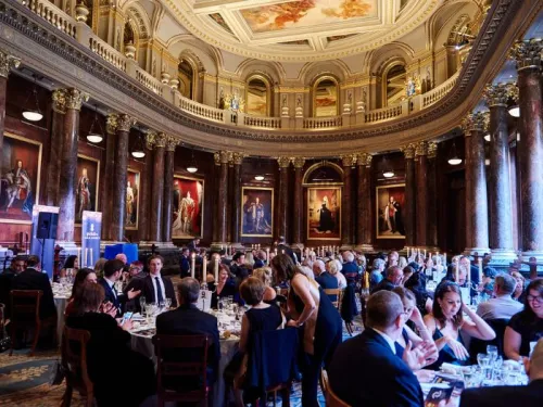 Tables of people in black tie attire in a large, ornate dining room as part of a gala event