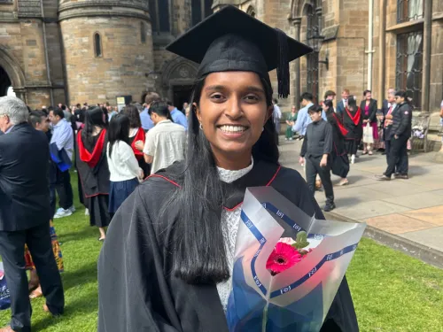 Shaumya wearing a graduation robe and cap, smiling at the camera and holding flowers.