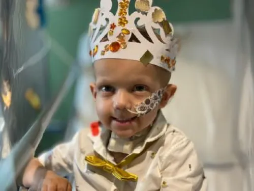 A young boy sitting on a hospital bed, with a tube in his nose, wearing a paper crown with craft jewels on it, and smiling