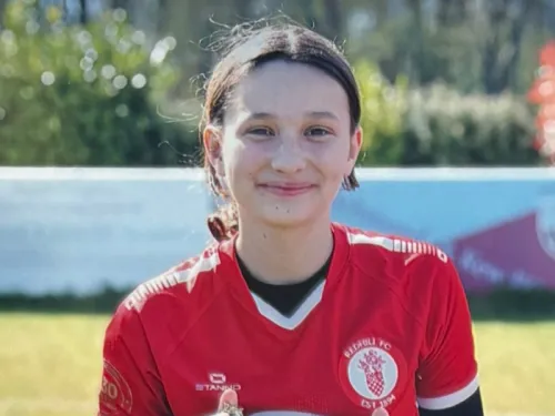 A young girl in her red football kit smiling at the camera in a park