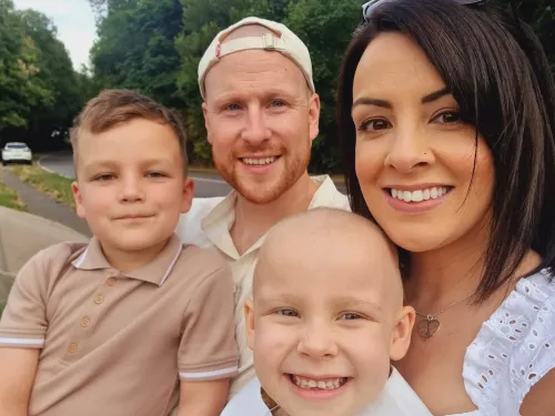 Woman smiling and facing camera with her husband and two children. One boy and one girl. The little girls has no hair and has cancer 