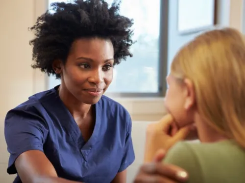 A black nurse in a blue uniform touching the shoulder of a patient with a green top and brown hair 